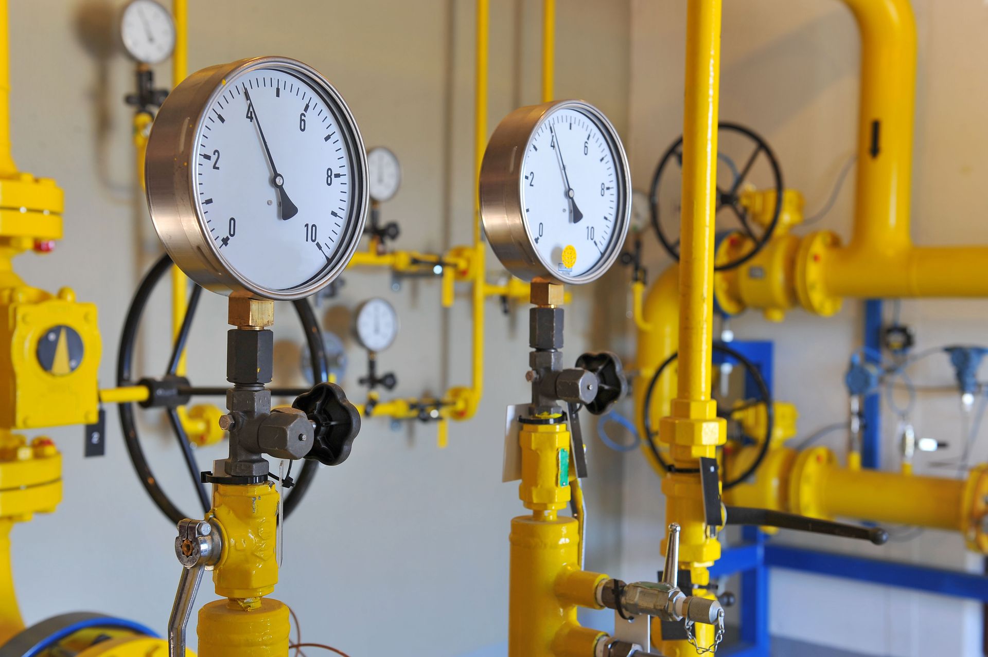Two circular analog pressure gauges mounted on yellow industrial pipes with valves and surrounding equipment.
