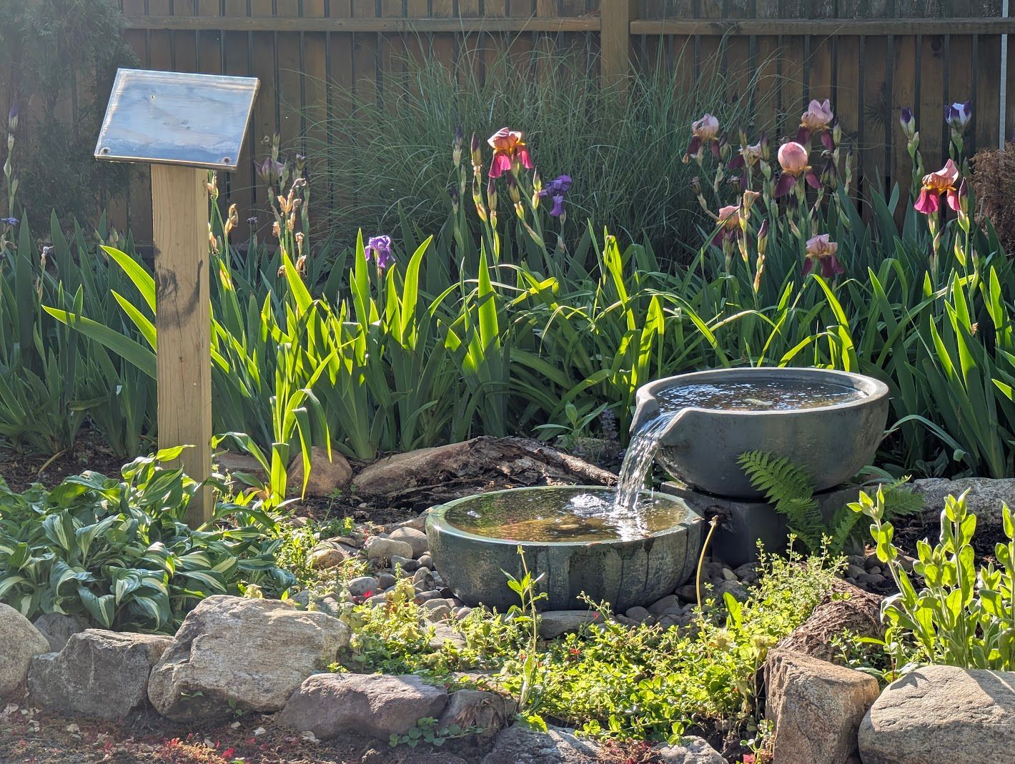 Garden with a small water fountain, flowers, and a sign post. Sunlight illuminates the scene.