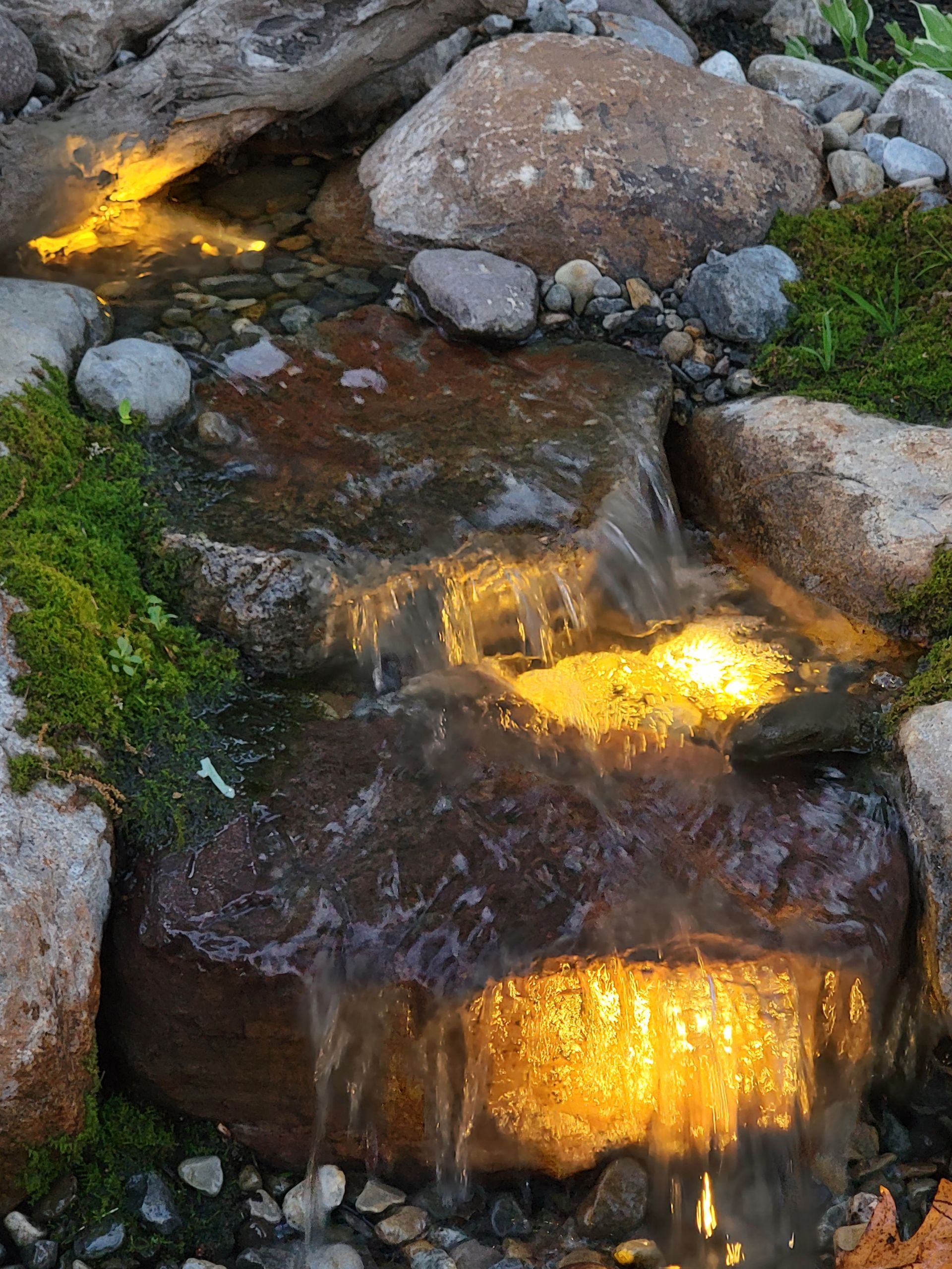 Water cascading over rocks in a small illuminated waterfall with moss and stones.