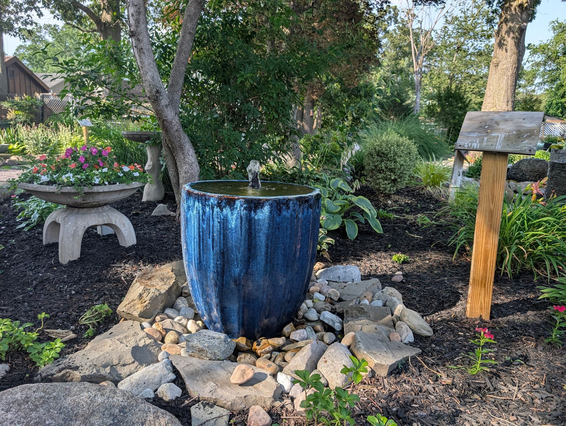 Blue glazed water fountain in a garden, surrounded by rocks and dark mulch, with a signpost and flowers.