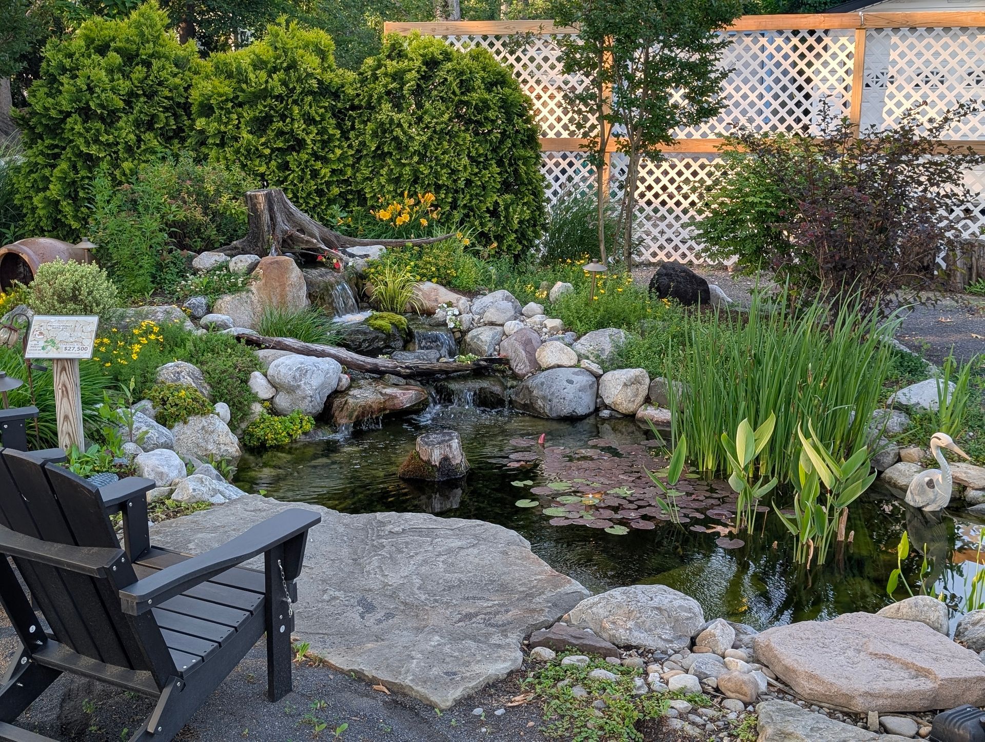 Pond with a small waterfall, surrounded by rocks and lush plants; an Adirondack chair sits nearby.