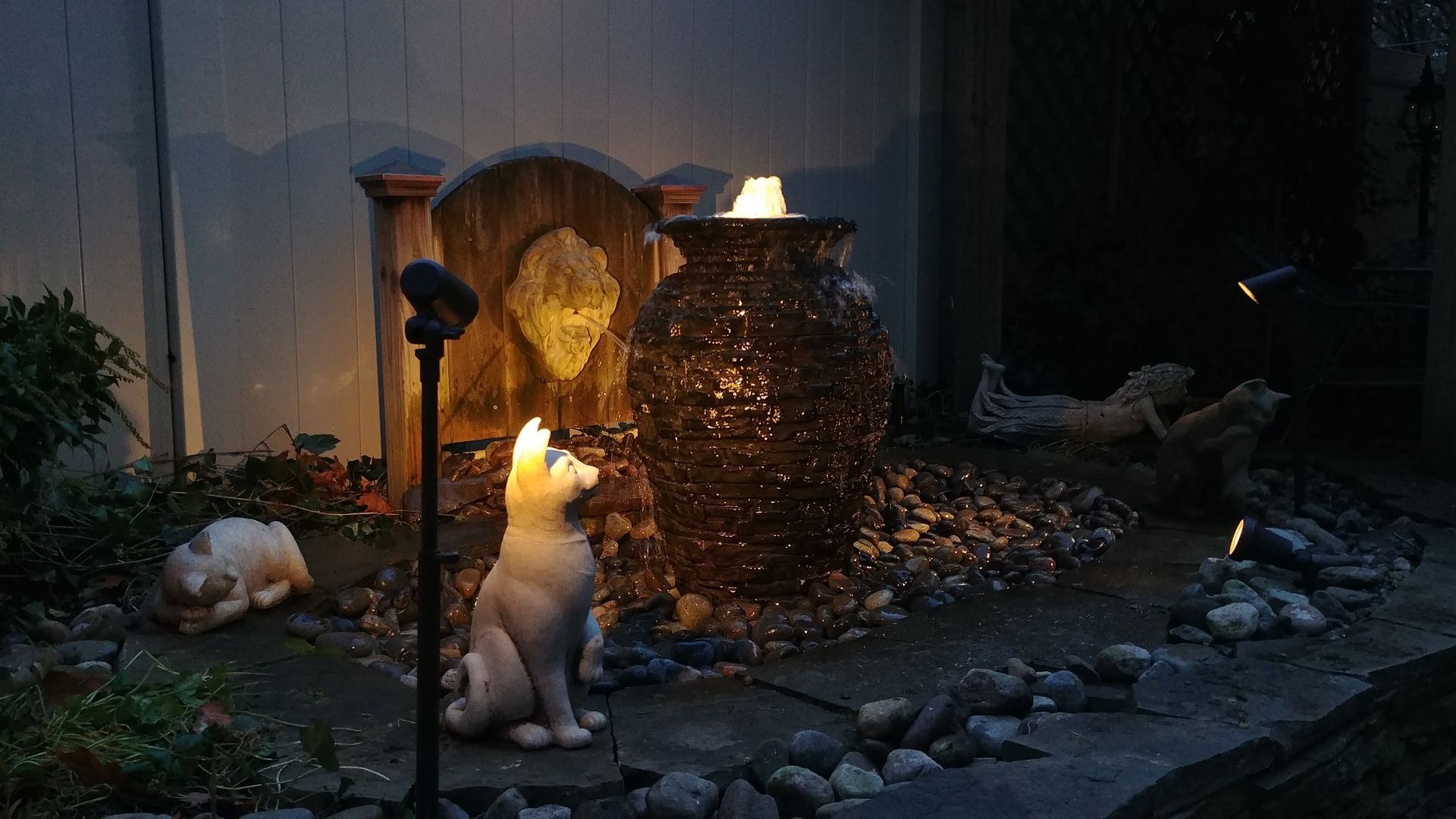 A night scene with a fountain, lit by spotlights, features stone statues of a dog and rabbit in a garden setting.