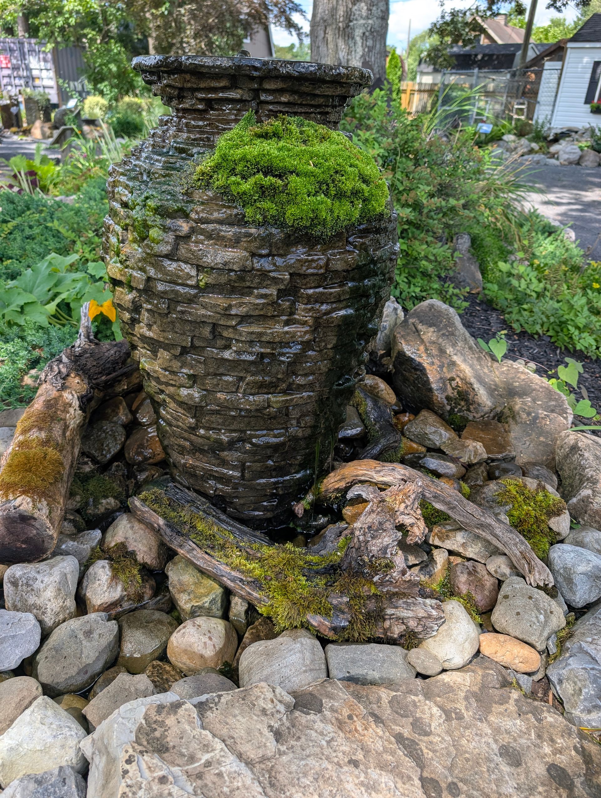 Water fountain made of stacked stone with greenery, surrounded by rocks and plants outdoors.