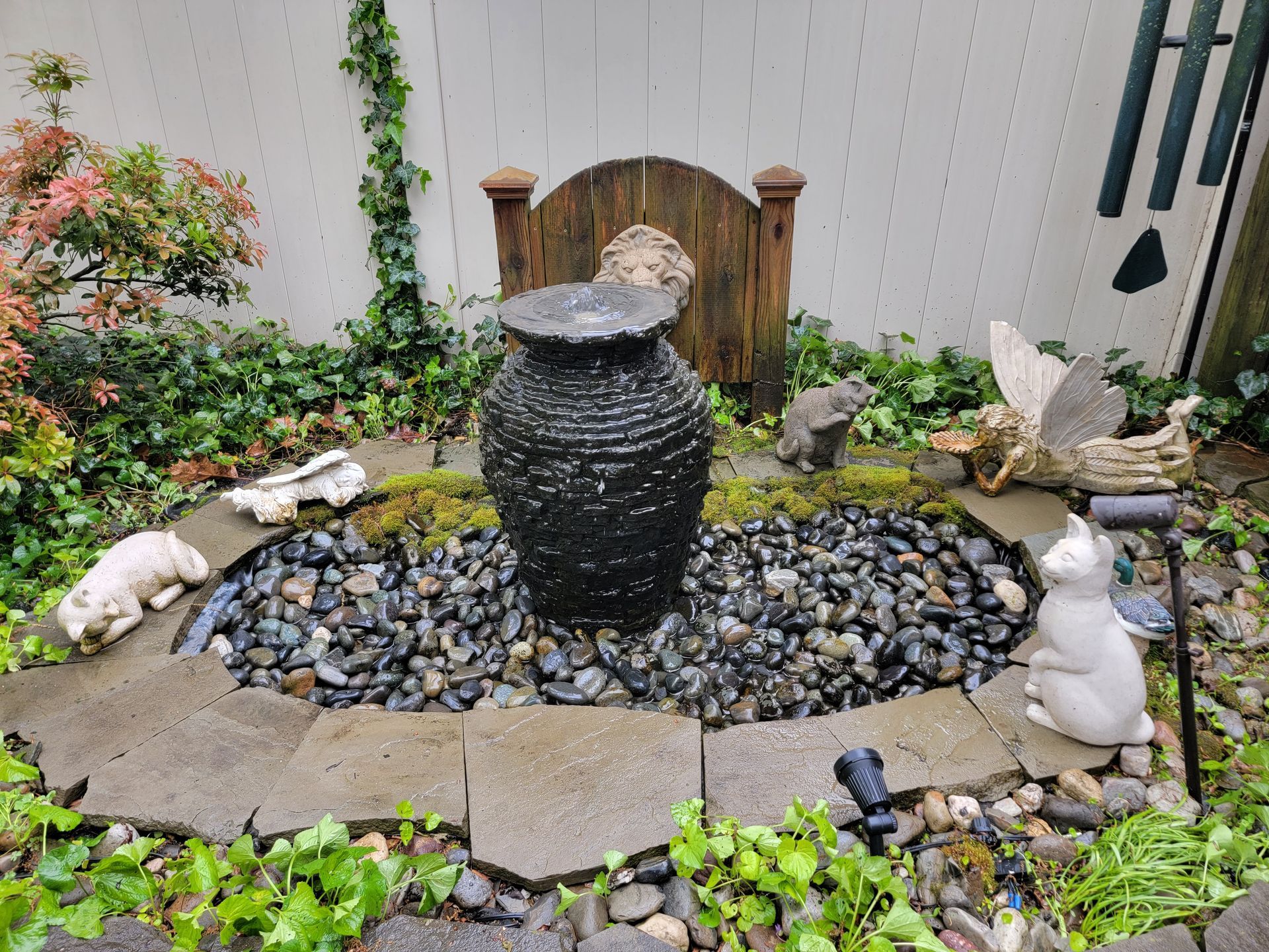Small decorative fountain with black rocks and stone edging; cat and squirrel statues; ivy, green plants.