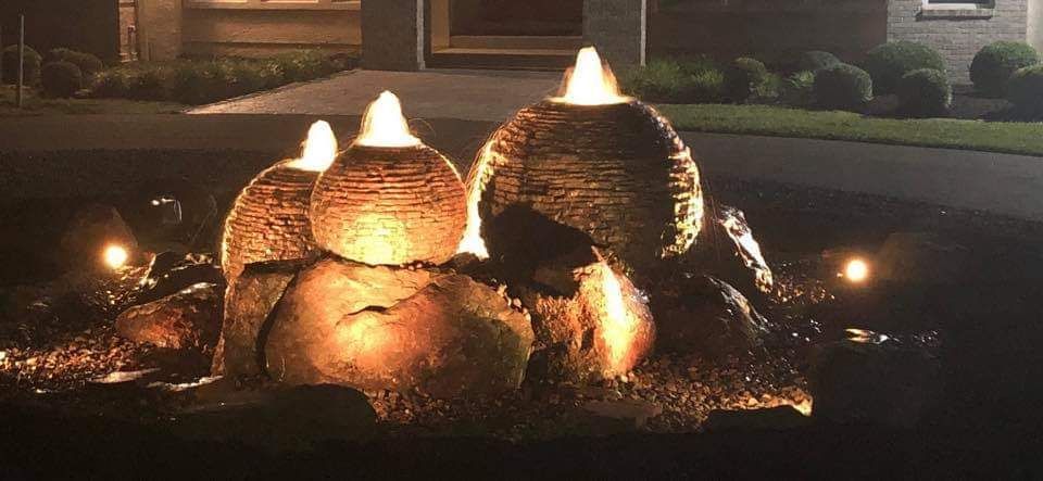 Lit outdoor water fountain, with large textured spheres and surrounding rocks, at night.