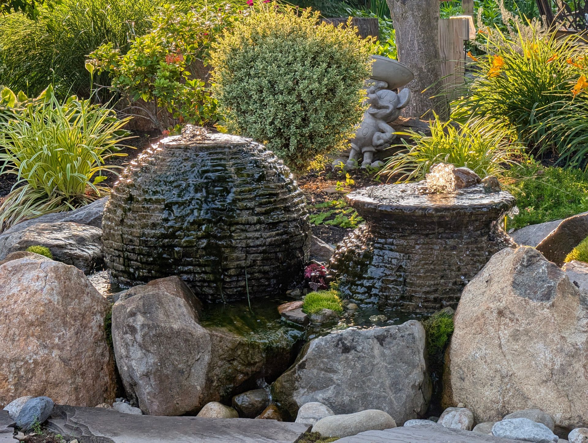 Water fountain with a stone globe and pot, surrounded by rocks and greenery in a garden.