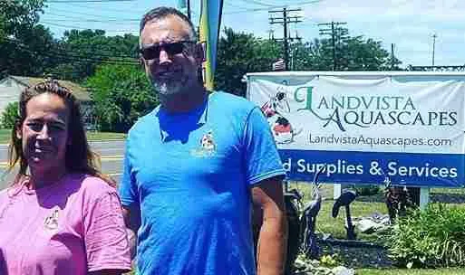 Two people stand near a sign for Landvista Aquascapes. Man wears blue, woman wears pink. Outdoors, sunny.