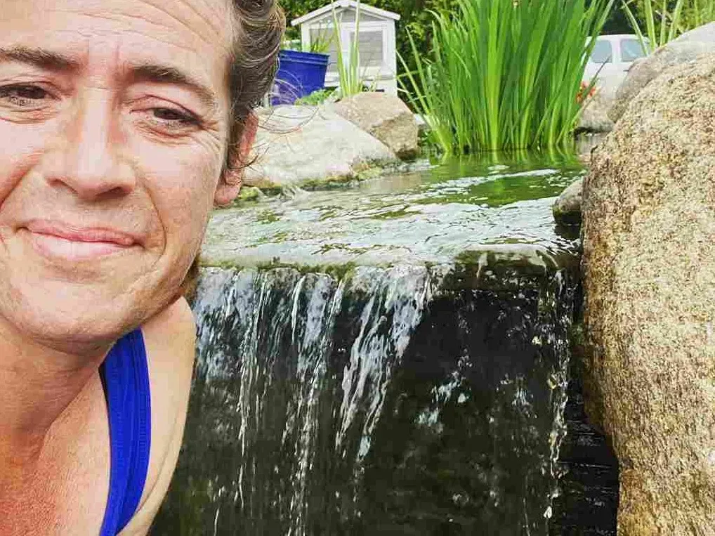 Person smiling near a waterfall flowing into a pond. Green plants and large rocks are visible.