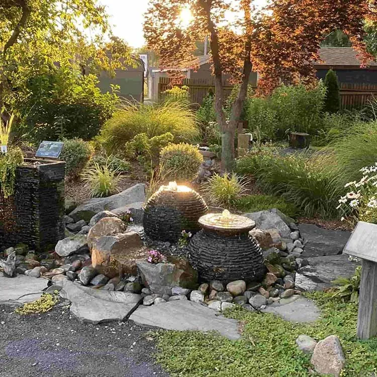 A garden with water fountains of round, textured black pots on a bed of rocks, illuminated by sunlight.