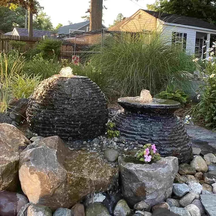 Water fountain with two dark, textured stone bowls in a garden setting. Water cascades into a pool.
