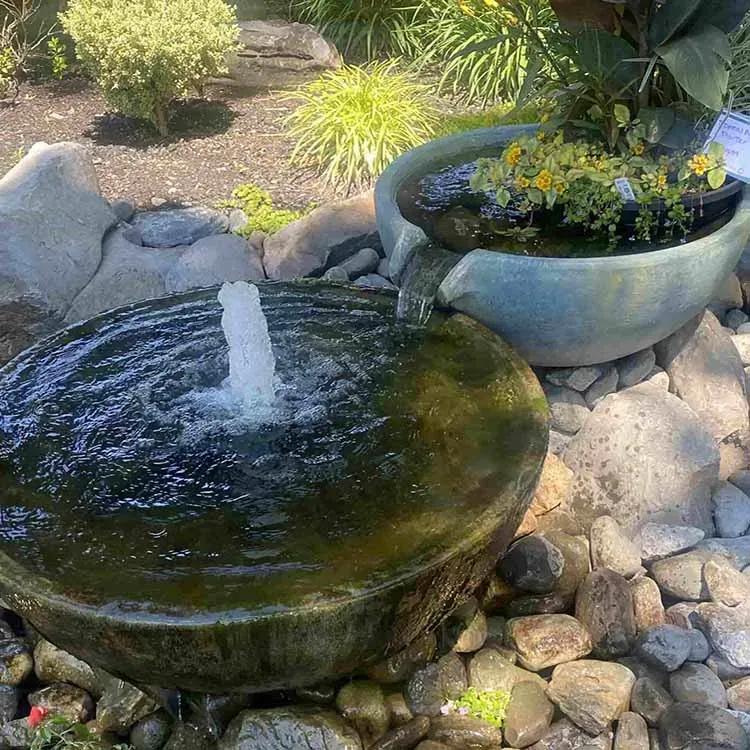 Water fountain with two stone bowls, overflowing with water, surrounded by rocks and plants.