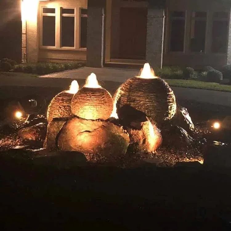 A nighttime view of a lit water fountain in front of a house. The fountain features three glowing orbs.