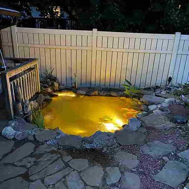 Lit-up backyard pond surrounded by rocks and a white fence at night.
