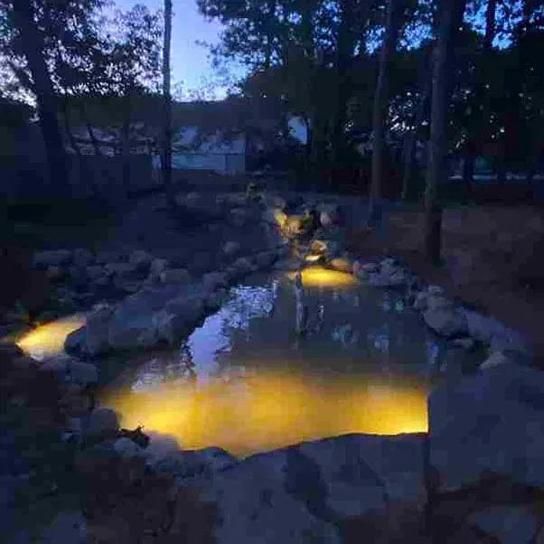 Night view of illuminated water feature with rocks, trees, and golden lights in a backyard.