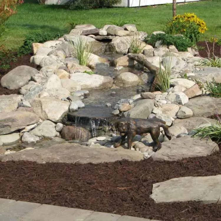 Water feature with waterfall and wolf statue. Rocks, plants, brown mulch. Sunny outdoor setting.