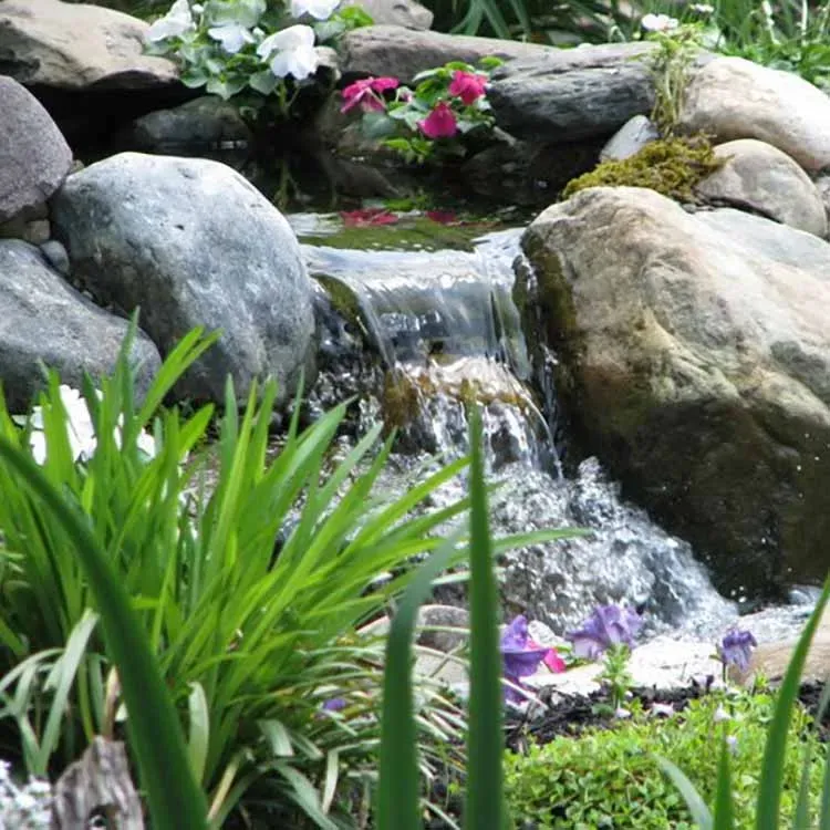 Small waterfall cascading over rocks, surrounded by green plants and colorful flowers.