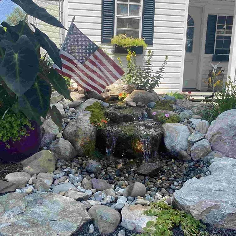 Water feature with waterfall, surrounded by rocks and plants, in front of a white house with an American flag.