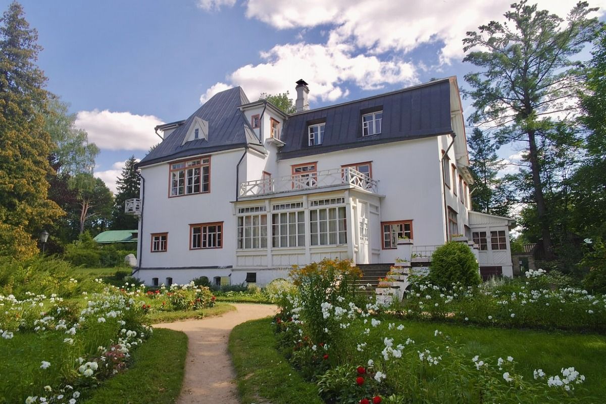A large white house with a black roof is surrounded by trees and flowers.