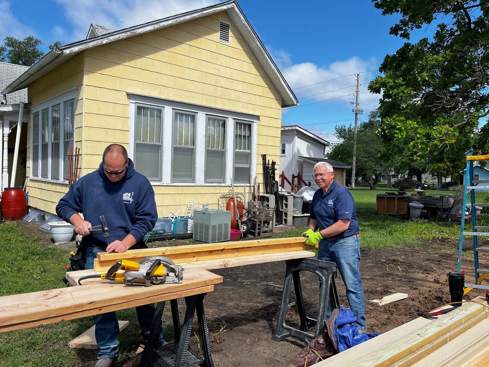 Two men are working on a wooden structure in front of a yellow house