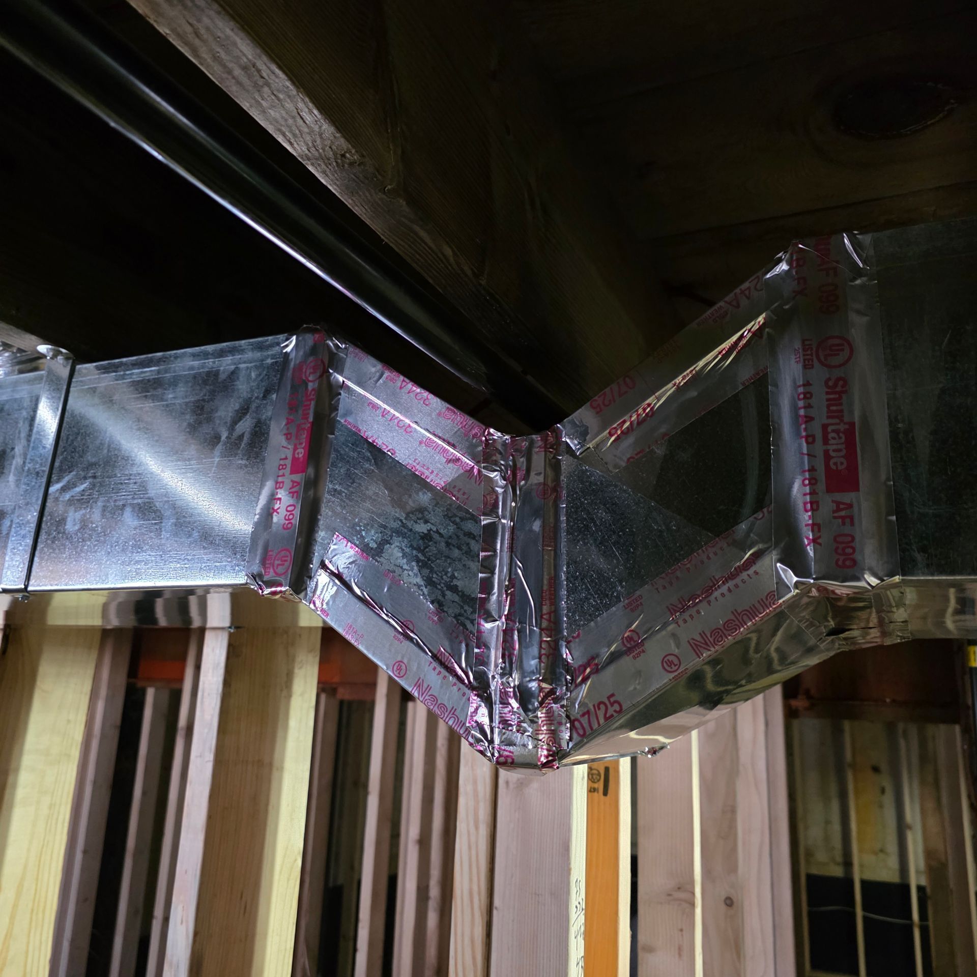 Metal HVAC ductwork with a zigzag bend running along a ceiling above unfinished walls.