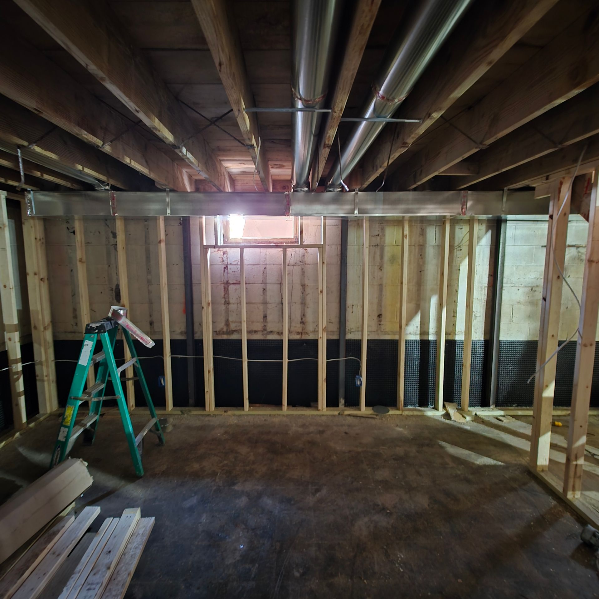 Partially framed basement with exposed ceiling joists, studs, a green ladder, and a small window.