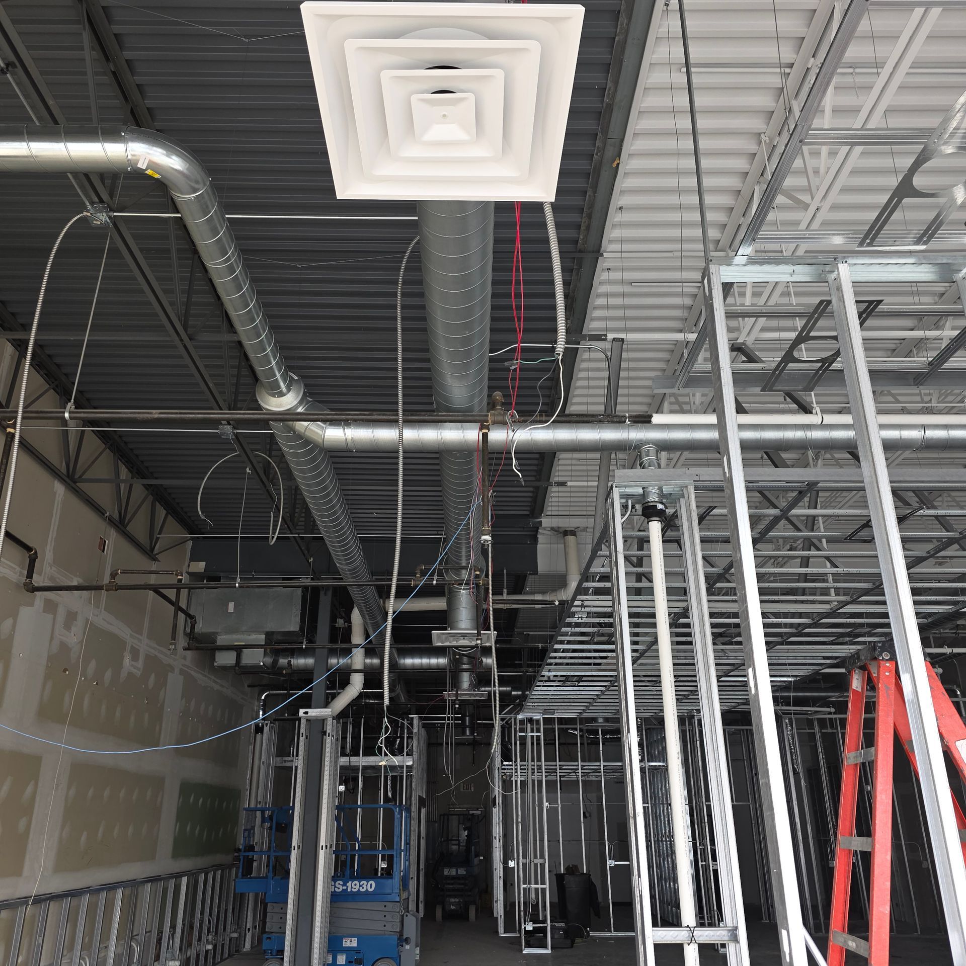 Indoor construction site with exposed ductwork, metal framing, and a blue lift beneath a white ceiling vent.