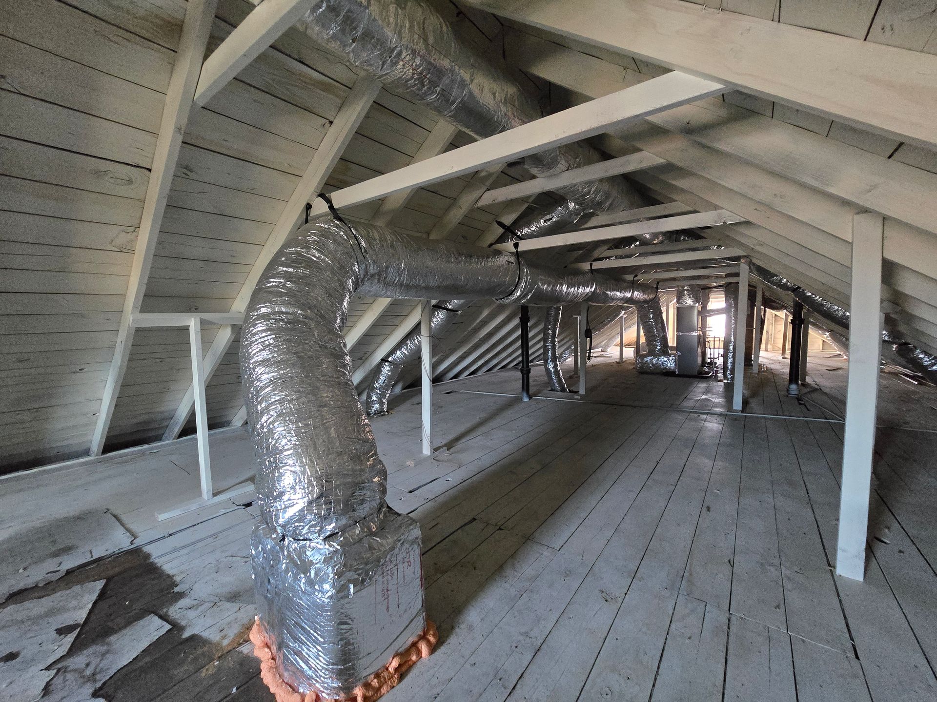 Attic with exposed silver HVAC ducting and white rafters over a wooden floor