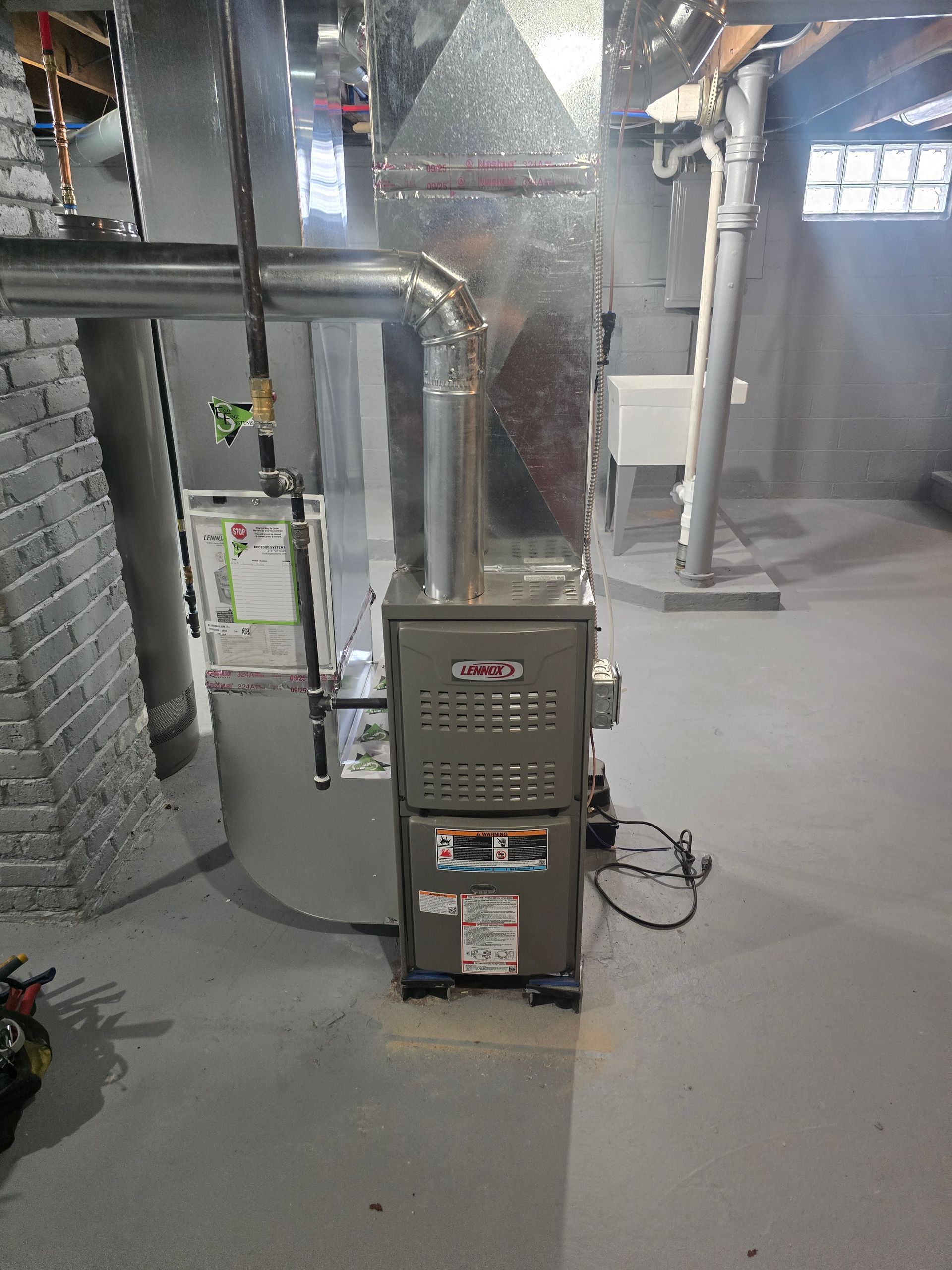 A grey HVAC furnace unit with metal ductwork stands on a concrete floor in a basement near a utility sink.
