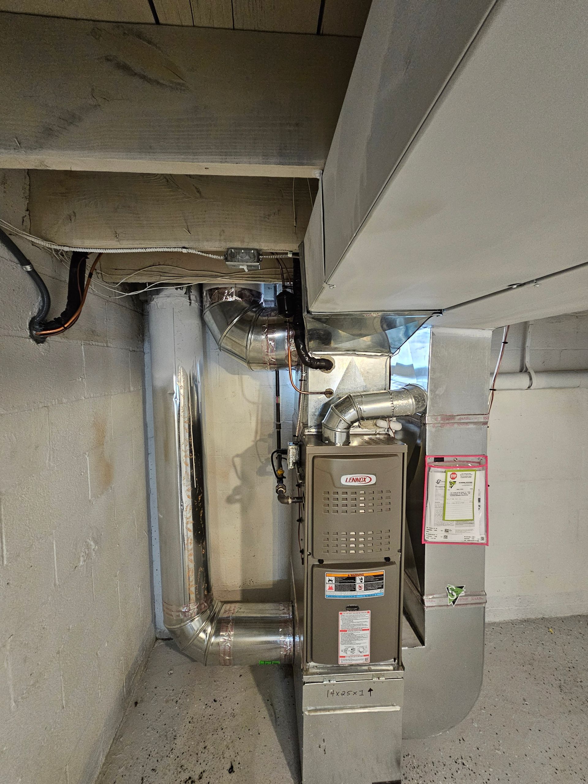 A furnace unit installed in a concrete basement, connected to silver metal ductwork under wooden ceiling joists.