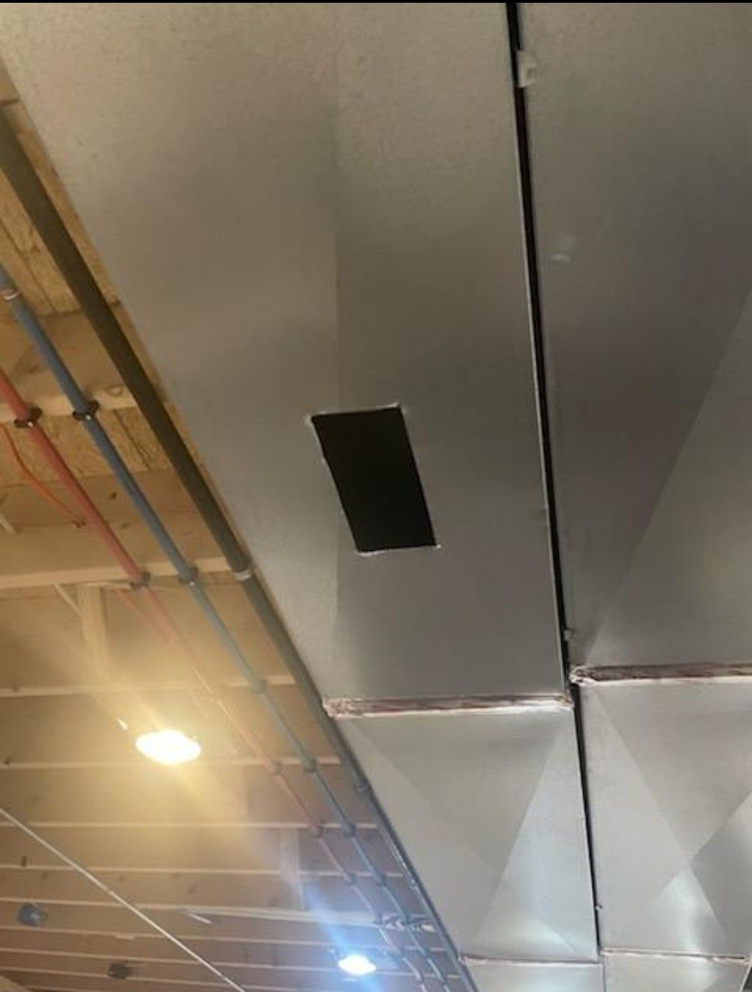 Rectangular black vent opening cut into a silver metal HVAC duct suspended from a wooden ceiling.