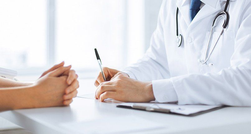 Doctor taking notes at desk, patient's hands clasped. Clinic setting, white coats.