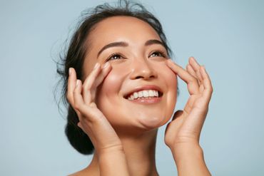 Smiling woman gently touching her face against a light blue background.