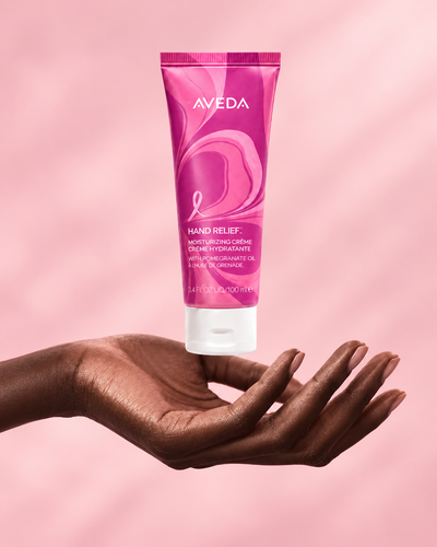 Pink Aveda tube floating above an outstretched hand on a soft pink background