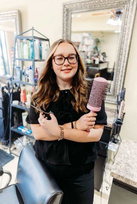 Woman in a salon holding a purple hairbrush, standing beside a mirror and styling station.