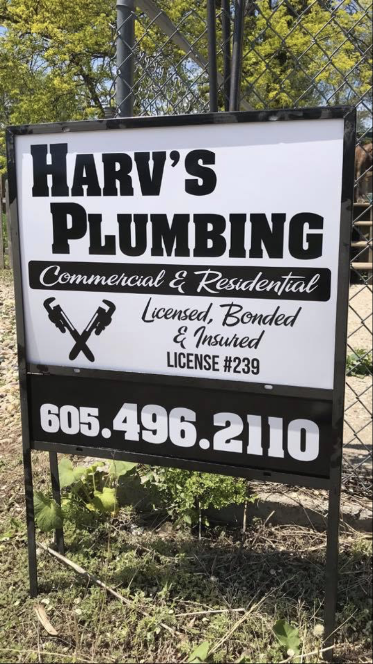 A harv's plumbing sign is sitting in the grass next to a chain link fence.