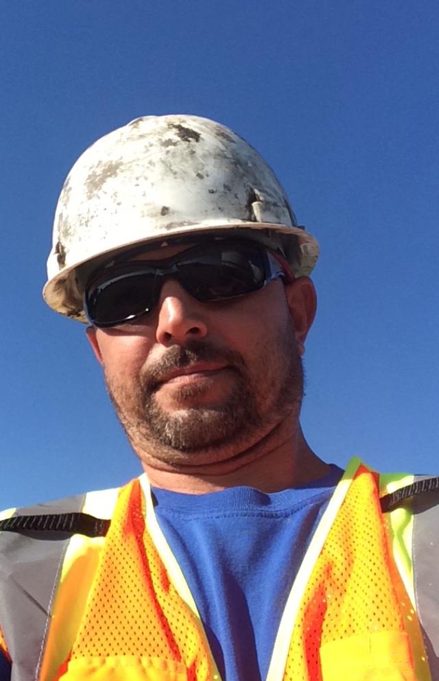 A man wearing a hard hat and sunglasses is standing in front of a blue sky.