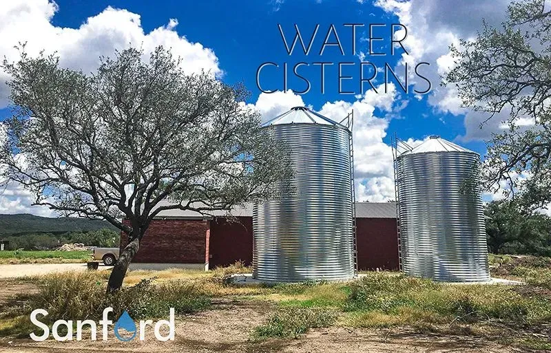 Two large, corrugated metal water cisterns stand outside a red building under a blue sky, text reads 