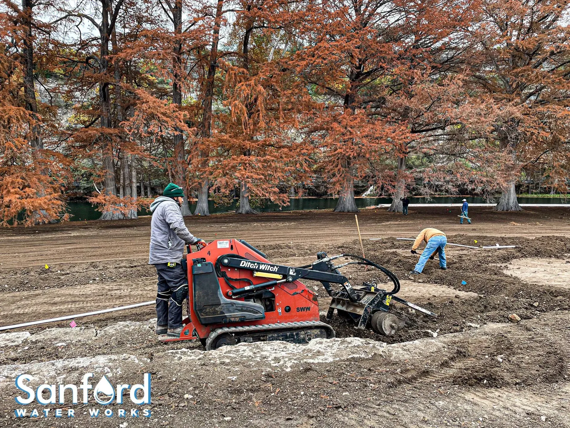Construction workers operating machinery in a field with brown soil and fall foliage in the background.