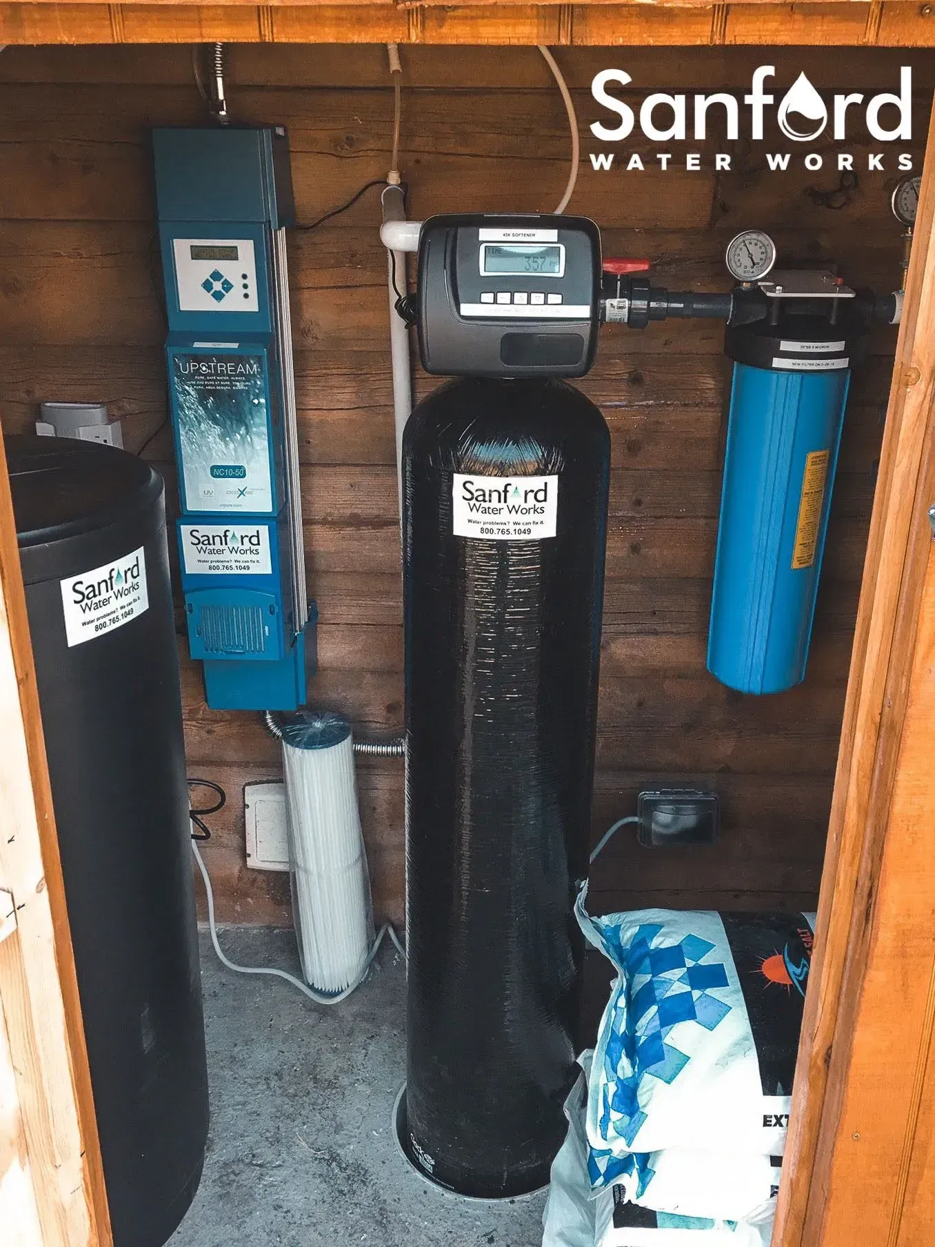 Water treatment system in wooden enclosure with Sanford Water Works branding. Black, blue, and white equipment.