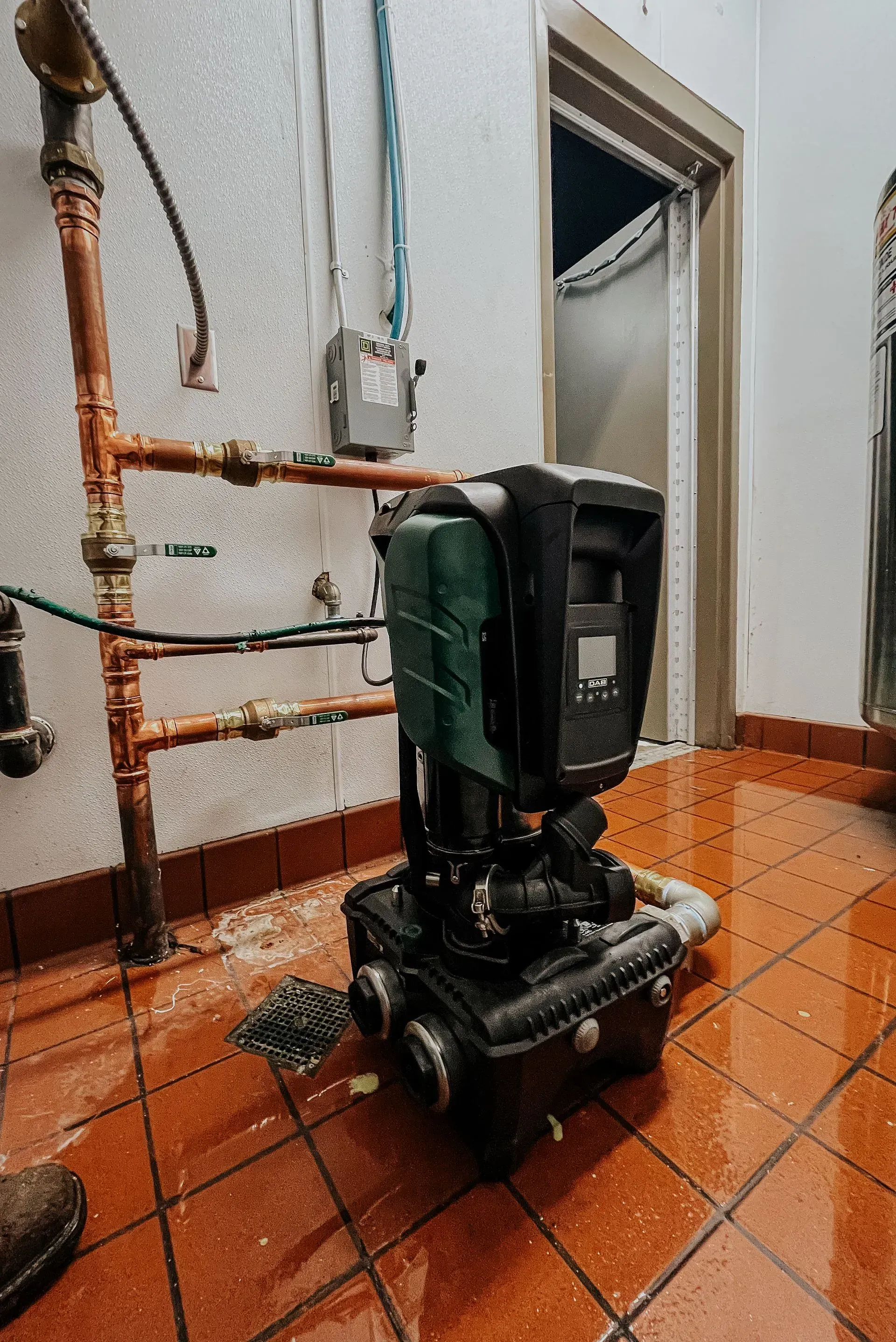 Water pump on wet, tiled floor next to copper pipes and a doorway.