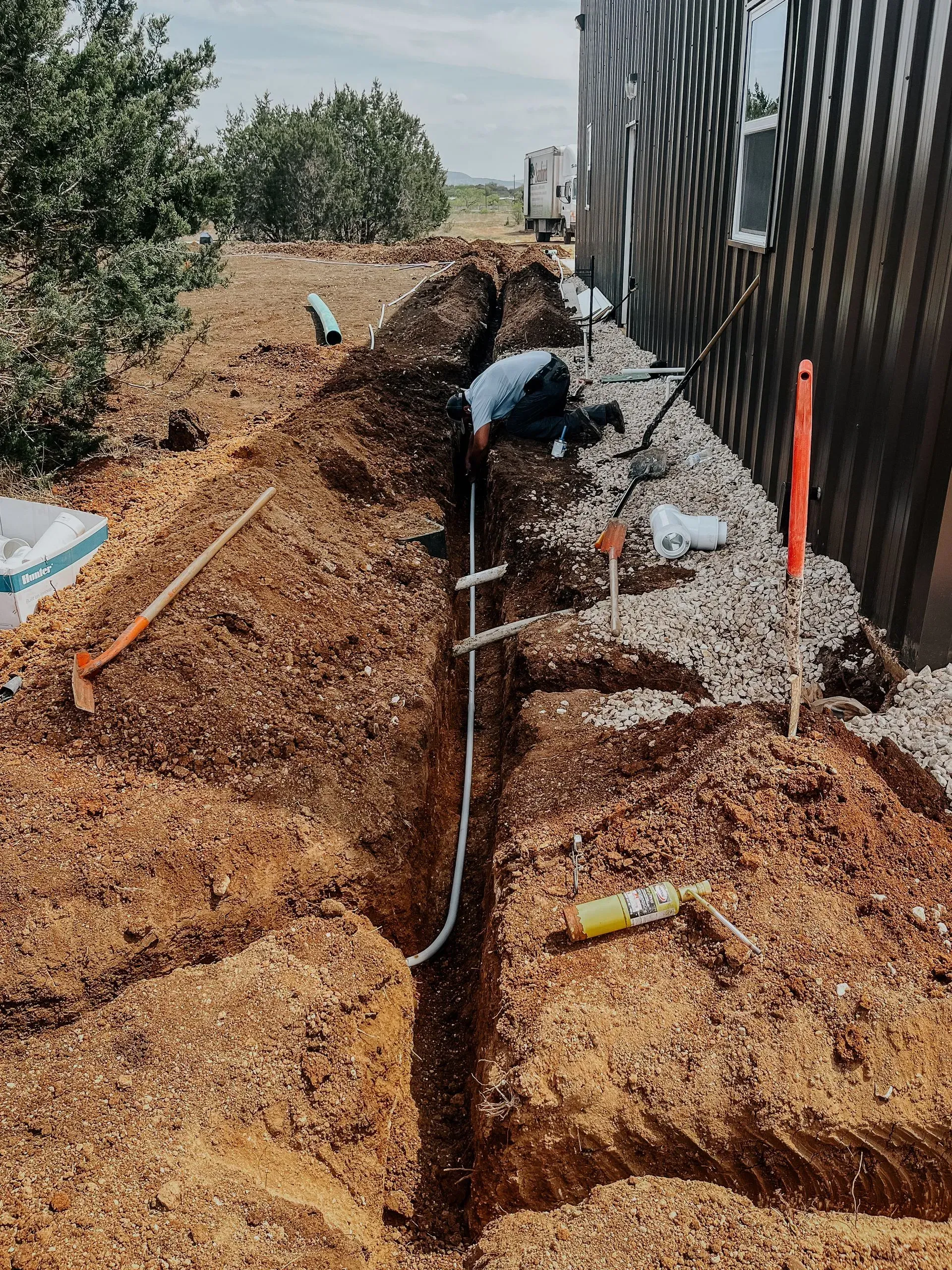 Person working in a trench, laying pipes. Brown soil, gray pipes, and a building are visible outdoors.