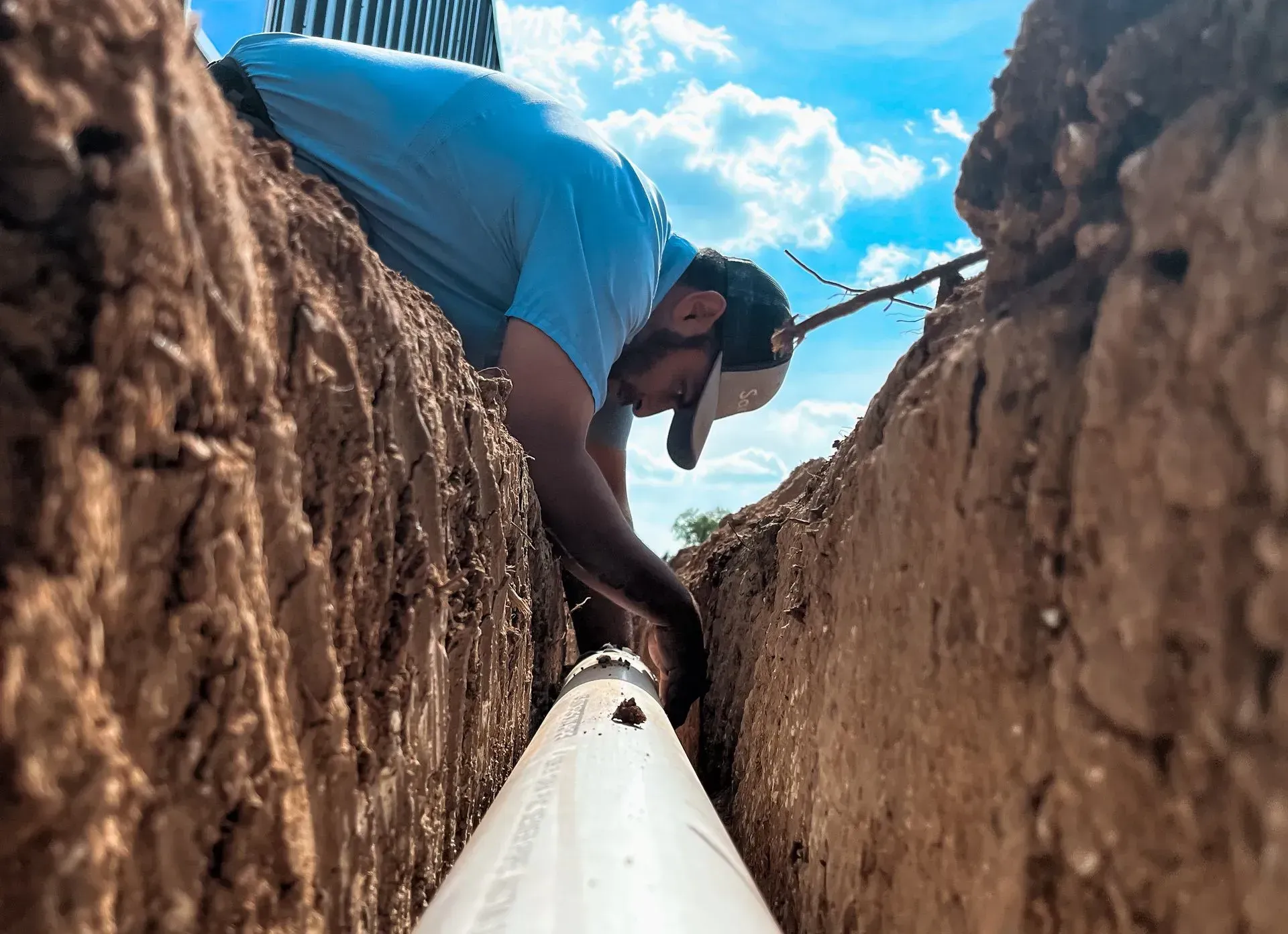 Man in a trench installing pipe under a partly cloudy sky.