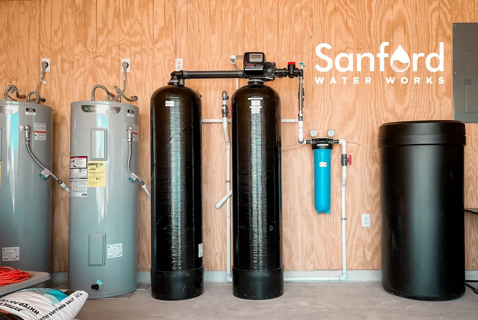 Water filtration system with two black tanks, blue filter, and water heaters against a wood-paneled wall.