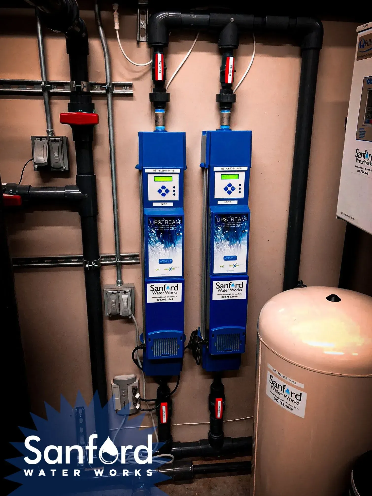 Two blue water filtration systems with pipes and a tank against a wall; Sanford Water Works logo.