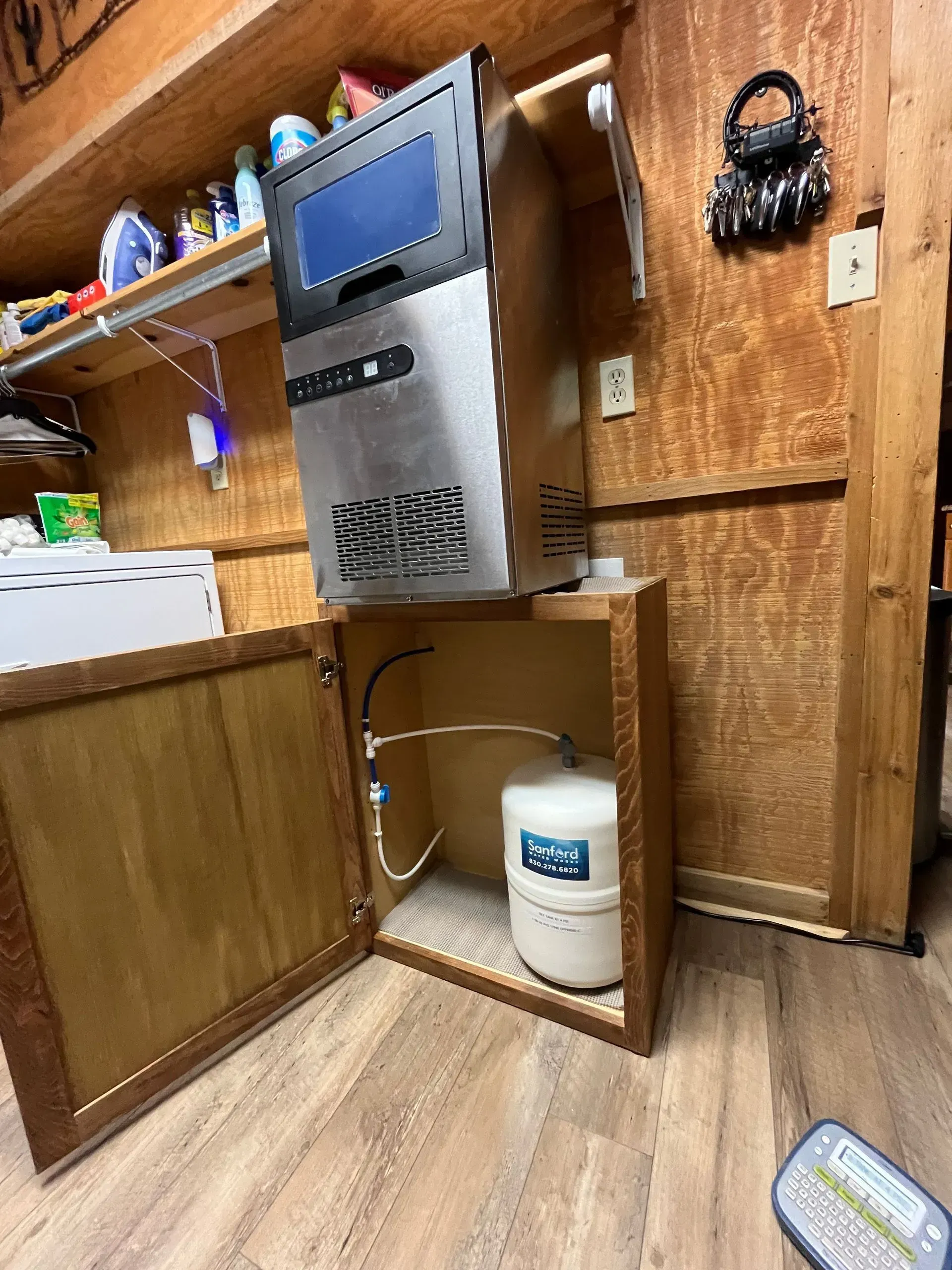 An ice machine mounted above a cabinet. The cabinet holds a water filter tank. Wooden walls.