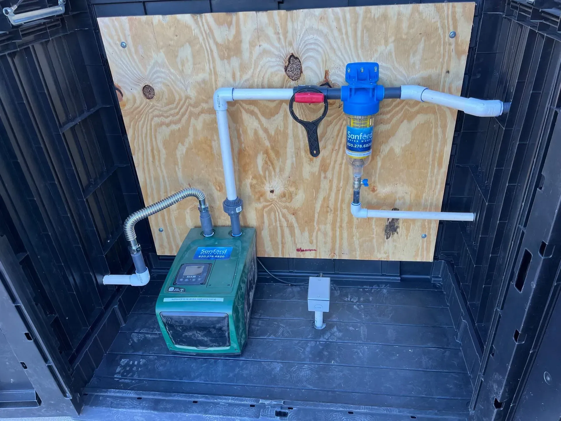 Water pump system mounted on plywood inside a black box, with white PVC pipes and a blue filter.