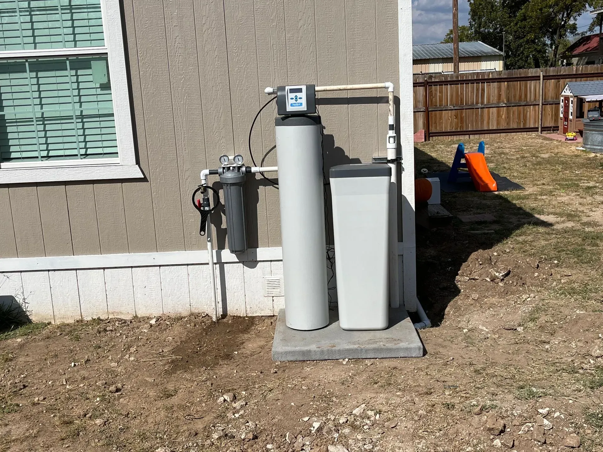 Water filtration system installed outdoors near a house with a window and a backyard with a slide.