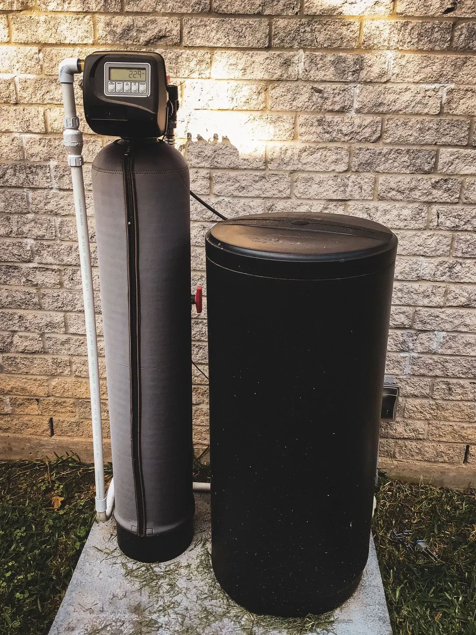 Water softener system, a tall black tank next to a cylindrical salt tank, stands outside against a brick wall.