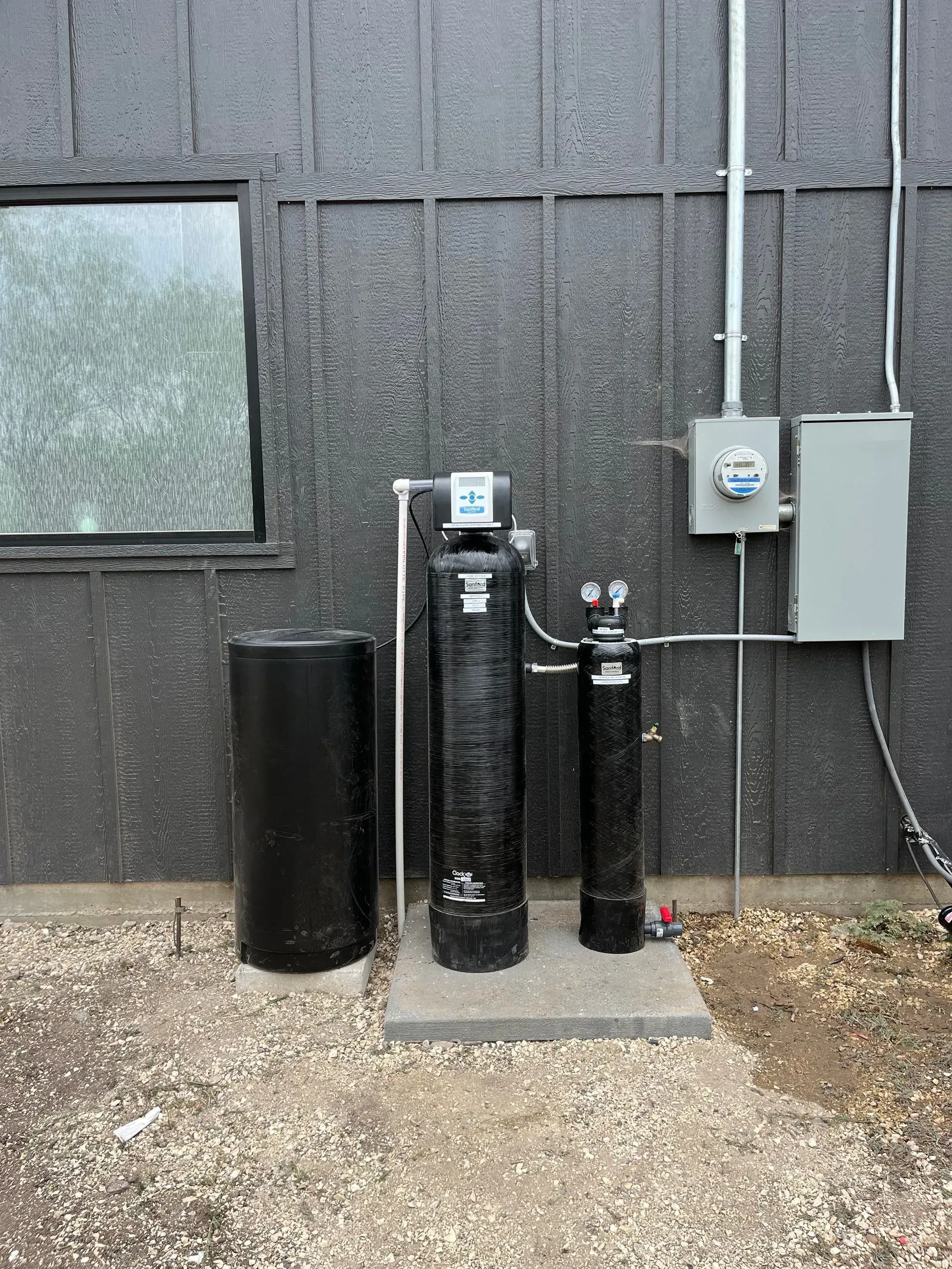 Water filtration system and electrical box against a dark exterior wall, gravel ground.