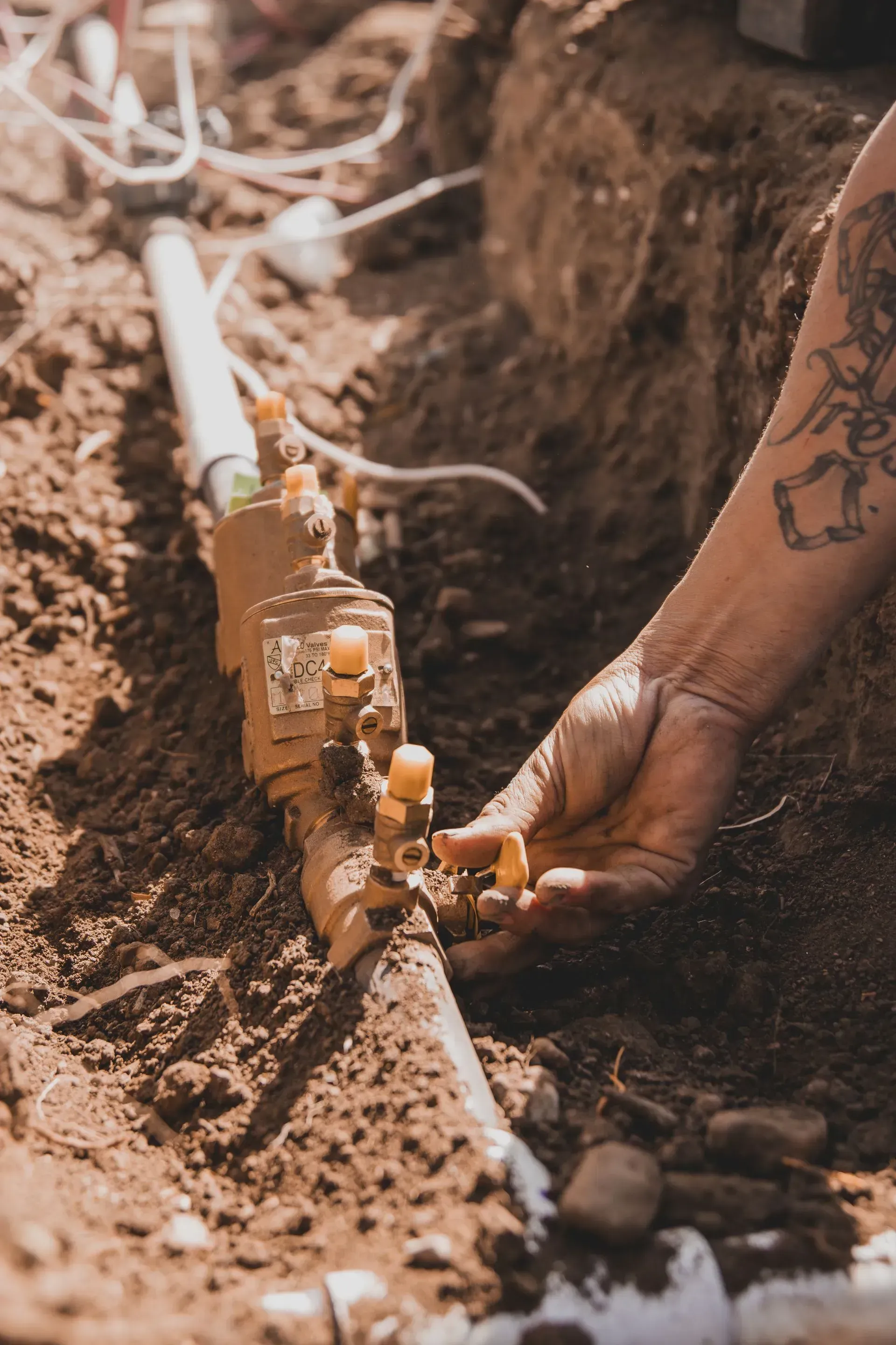 Person working on buried pipes, adjusting valves. Brown dirt surrounds copper-colored plumbing and white plastic.